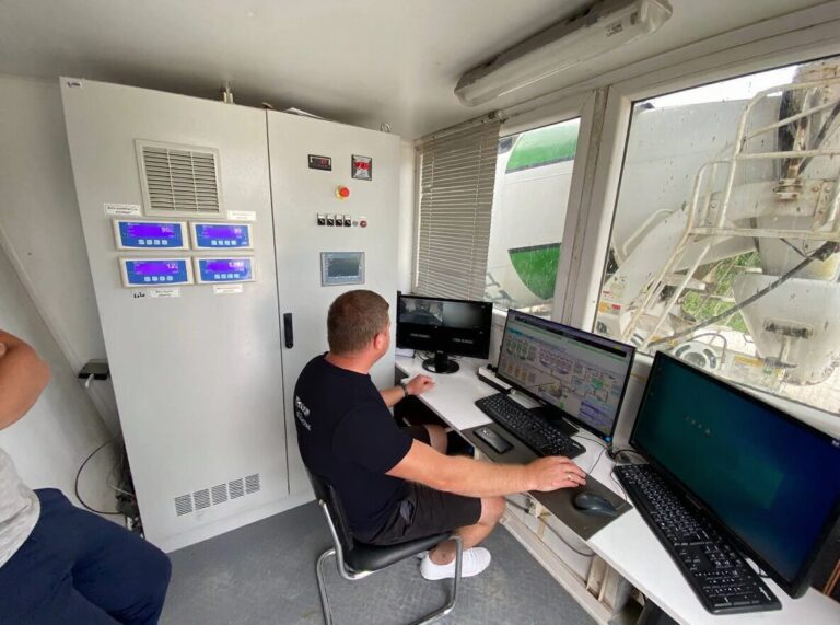 Operator working with multiple computer monitors and control panels inside a control room, demonstrating Concrete batching plant automation in action.